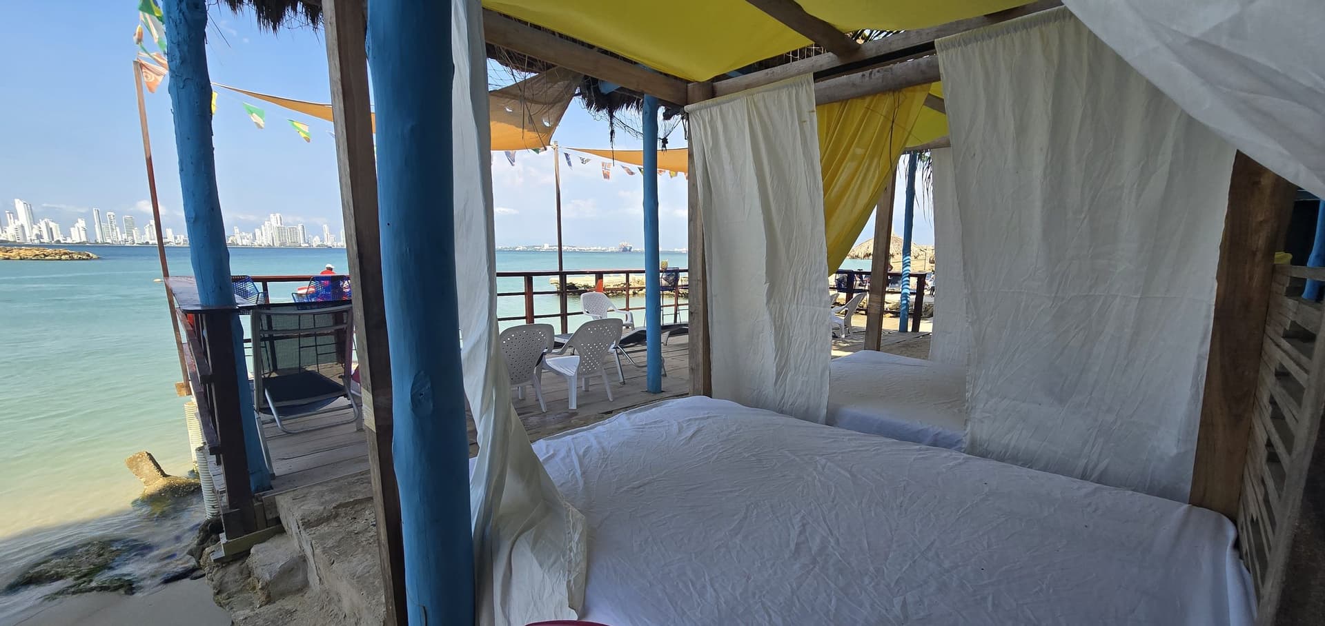 Cama de playa bajo techo con cortinas blancas y amarillas, junto al deck y frente al mar.
