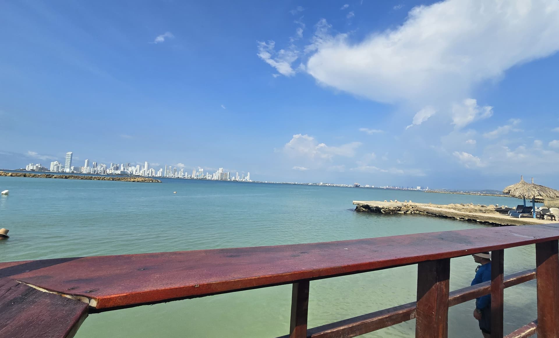 Baranda de madera de la terraza frente al mar, con el espolón y la ciudad en el horizonte.